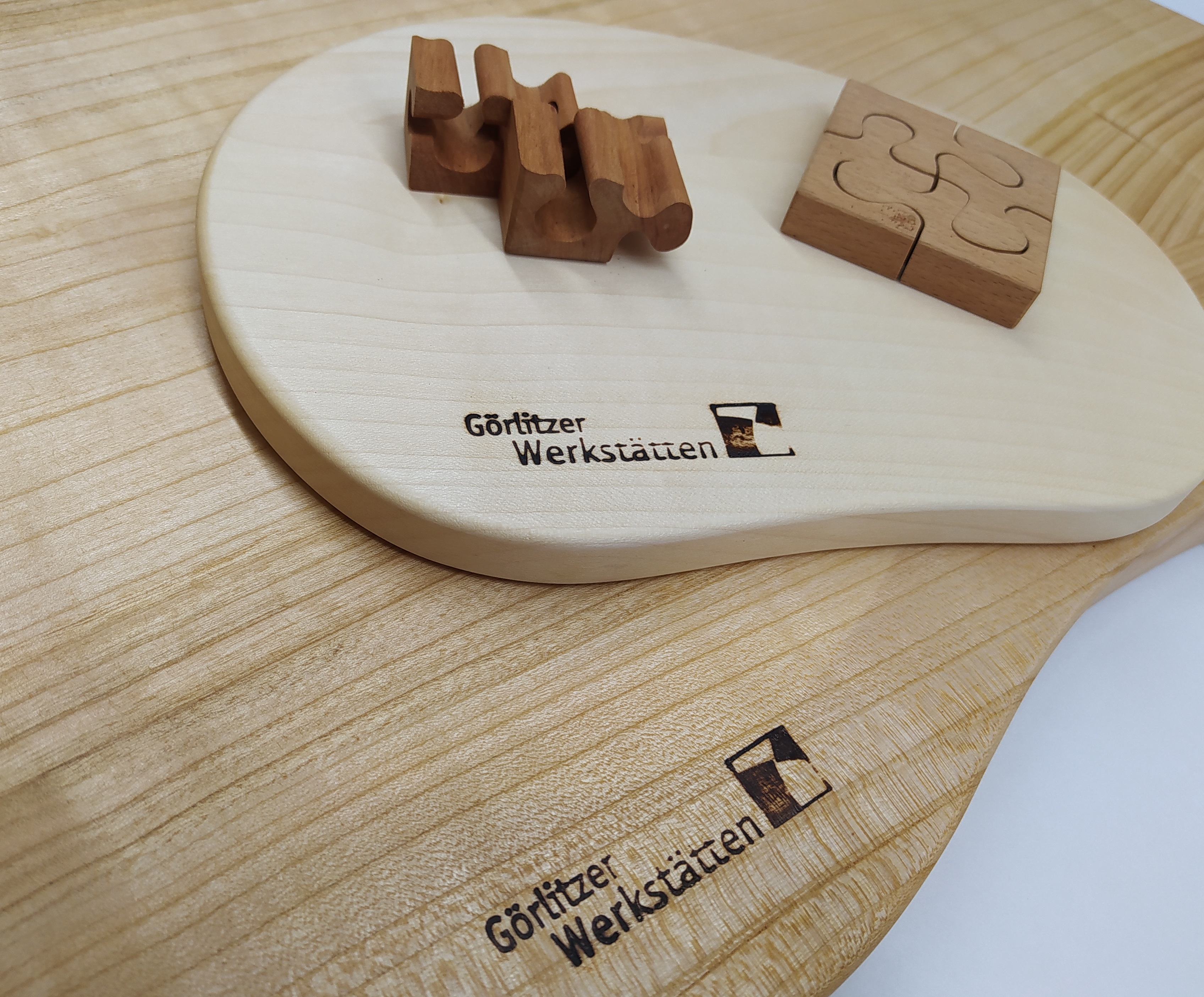 Ein kleines geschwungenes Holzschneidebrett liegt auf einem größeren geschwungenen Holzschneidebrett. Auf dem keinem Holzschneidebrett liegen zwei aus dem Holz der Schwarzerle bestehende 3D-Puzzel. Ein 3D-Puzzel ist davon in seine Einzelteile zerlegt.