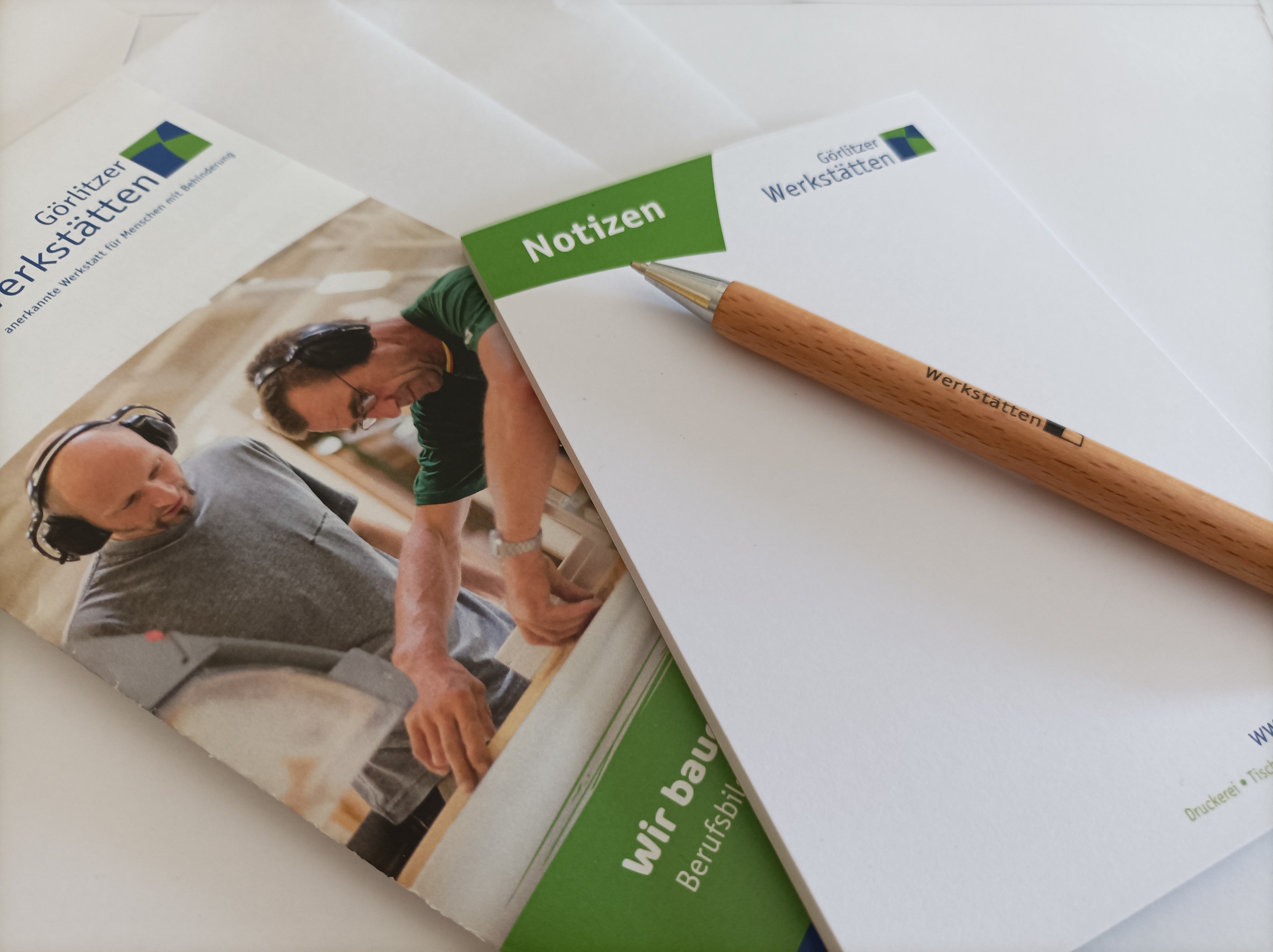 : Übereinandergestapelt liegen etwas leicht versetzt auf einem Flyer der Görlitzer Werkstatt ein Notizblock aus weißem Papier, auf dem Papier befindet sich in der linken oberen Ecke ein grünes Feld mit dem Wort Notizen, daneben ist das Logo der Görlitzer 