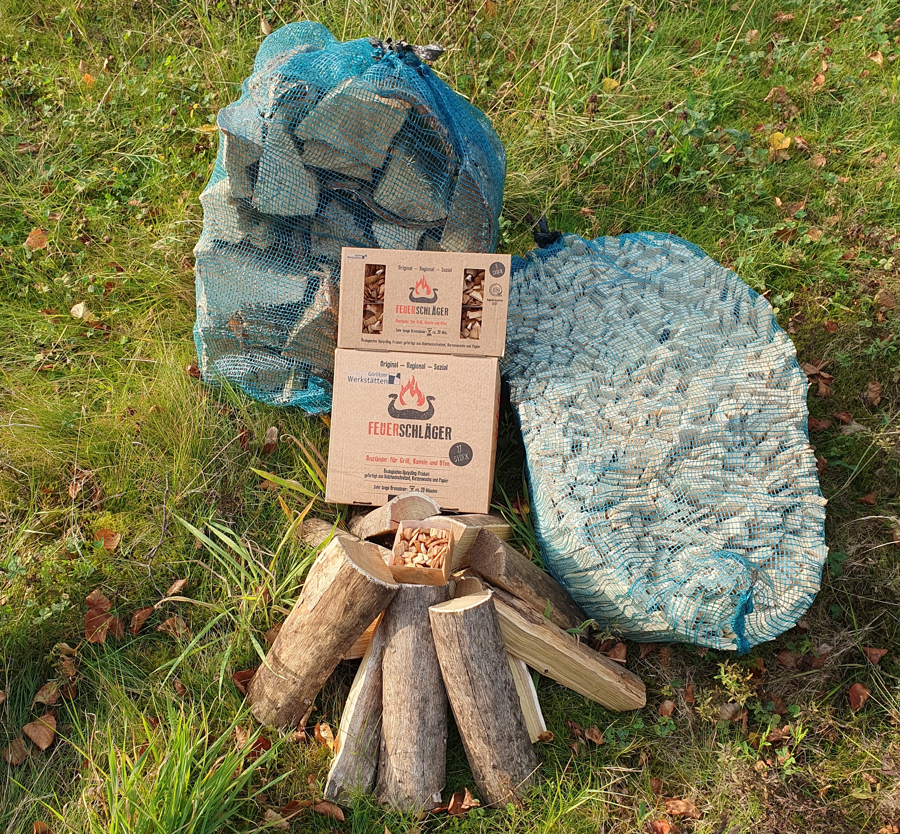 Auf einer grünen Wiese liegen zwei Packungen Grillanzünder der Marke Feuerschläger. Um die Grillanzünder herum ist ein Sack Anzündholz, ein Sack Kaminholz und ein Scheit Feuerholz angeordnet.
